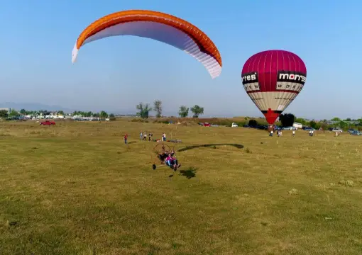 Motor paraglider with an orange wing taking off from a grassy airfield