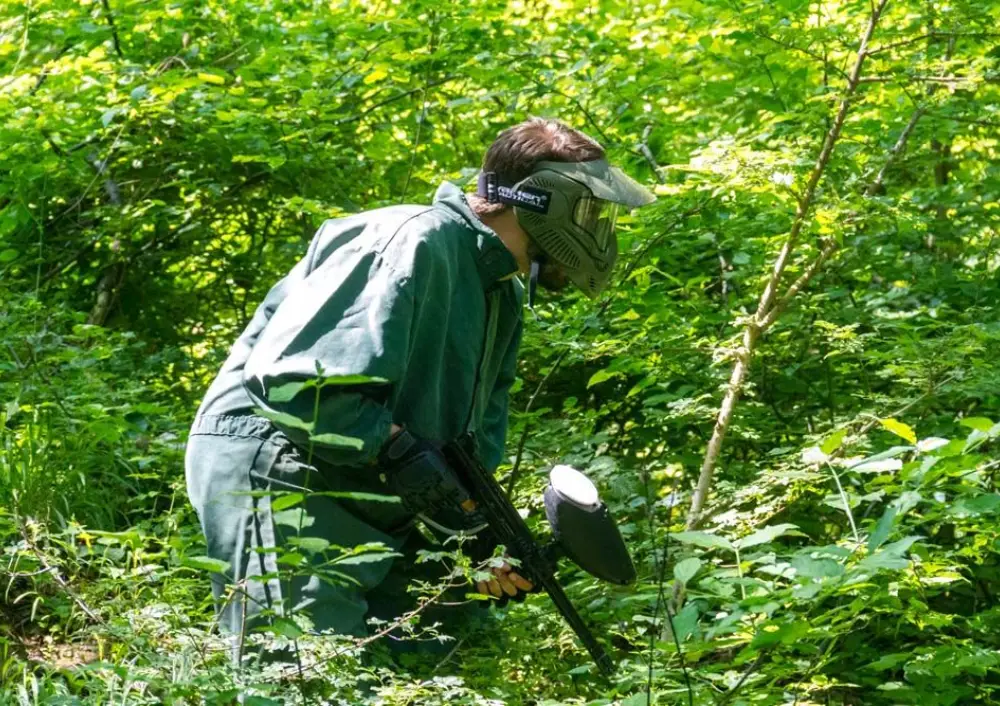 Crouching paintball player aiming through thick green undergrowth